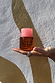 NFD Eau De Parfum 002 amber glass bottle with a pink marbled cap, held in a palm against a textured gold wall.