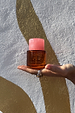 NFD Eau De Parfum 002 amber glass bottle with a pink marbled cap, held in a palm against a textured gold wall.
