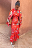 Thumbnail for Tomato Tapestry Frida Dress in red with sun motifs, layered with a coordinating cardigan and metallic shoes.