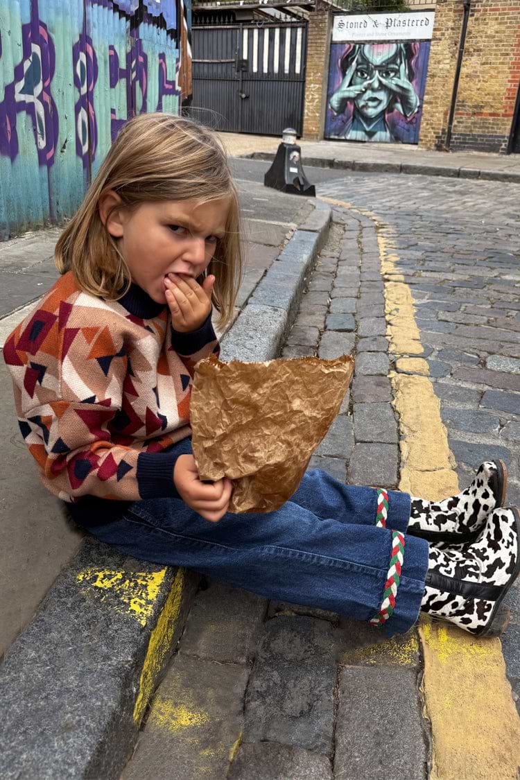 NFD Kids Aspen Jumper with a geometric knit pattern, worn with jeans and cow-print boots on a cobbled London street.