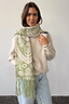 Olive Amara Knit Scarf in a green Mediterranean heart and floral print with tassels, layered over a fuzzy jumper.