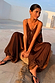 Chocolate Balloon Jumpsuit with a draped silhouette and gathered neckline, shown on a model sitting on a woven stool.