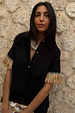 Black Raffia Shirt featuring tan raffia fringed sleeves and crochet shell-esque buttons against a stone wall.