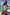 Thumbnail for Blue Starburst Bandeau Swimsuit with sun, palm and heart motifs and ruched side ties, worn by a pool.