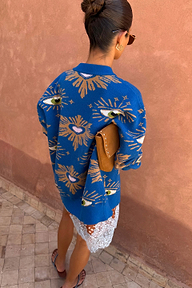 Blue Eye Cardigan with iconic eye and heart motifs, styled from behind with a white lace skirt and tan clutch.