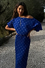 Blue Tilly Plisse Dress featuring silver dots and an off-the-shoulder neckline, worn by a model on a path.