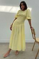 Thumbnail for Butter yellow Nara midaxi dress with a shirred bodice, puff sleeves and pleated skirt on a model leaning on a chair.