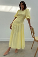 Butter yellow Nara midaxi dress with a shirred bodice, puff sleeves and pleated skirt on a model leaning on a chair.