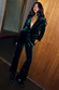Emerald Velvet Trousers and matching blazer with diamond studs on a model leaning against a wooden wall.