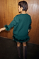 Bottle Green Jamie Jumper with detachable feather trims, shown from the back against wooden panelling.