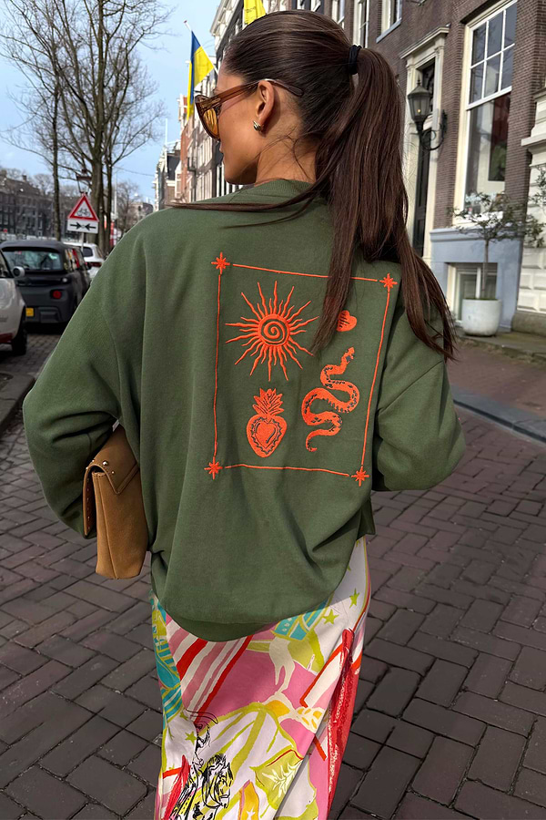 Khaki sweatshirt with orange sun and snake embroidery on the back, styled for a casual London look.