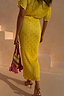 Sunshine Tilly Dress in yellow plissé with gold flecks and lettuce hem, held with a colourful beaded bag.