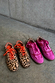 Pink and red leather trainers beside leopard print sneakers with orange laces on a grey concrete background.