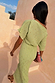 Pistachio Tilly Dress in green plisse with gold flecks, styled off-the-shoulder on a model walking up stairs.