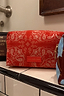 Thumbnail for Red Bandana Reversible Make Up Bag with an in-house designed paisley print and gold zipper on a tiled shelf.