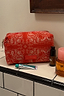 Thumbnail for Red and pink bandana print makeup bag with a gold zip, shown on a tiled bathroom counter next to skincare essentials.