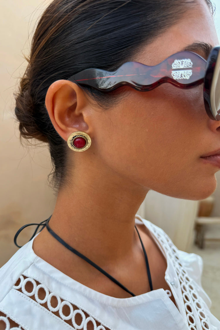 Deloris Red Circle Earrings in textured brushed brass with a red stone, shown on a model wearing tortoiseshell sunglasses.