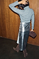 Silver Vegan Leather Jaspre Skirt with a grey knit and brown boots from behind against a wood wall.