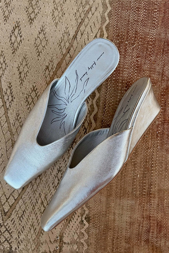 Silver metallic leather wedge mules featuring a pointed toe and sun-printed insole, resting on a patterned rug.