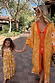 Girl's yellow cotton maxi sun dress with an orange sun print and shirred top, walking with a matching adult on vacation.