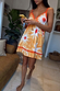Yellow Sun Mini Dress in cotton with an orange sun print, crochet trims and tie-front, worn by a woman indoors.