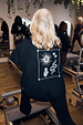 Black sweatshirt with ecru embroidery of a sun and snake, worn in a Pilates studio on a reformer.