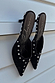 Black suede mules with silver stud detailing and scalloped edges, leaning against a white slatted background.