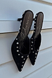Black suede mules with silver stud detailing and scalloped edges, leaning against a white slatted background.