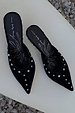 Pointed black suede mules with silver studs and scalloped edging, shown on a neutral stone background.