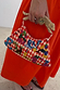 Red checkered Cirque Bag featuring hand-embroidered beads and a tiger-shaped wooden handle, held against an orange dress.