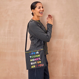 Charcoal Life is Tough Honey Drainage Bag with colourful text, worn crossbody by a smiling woman in a grey outfit.