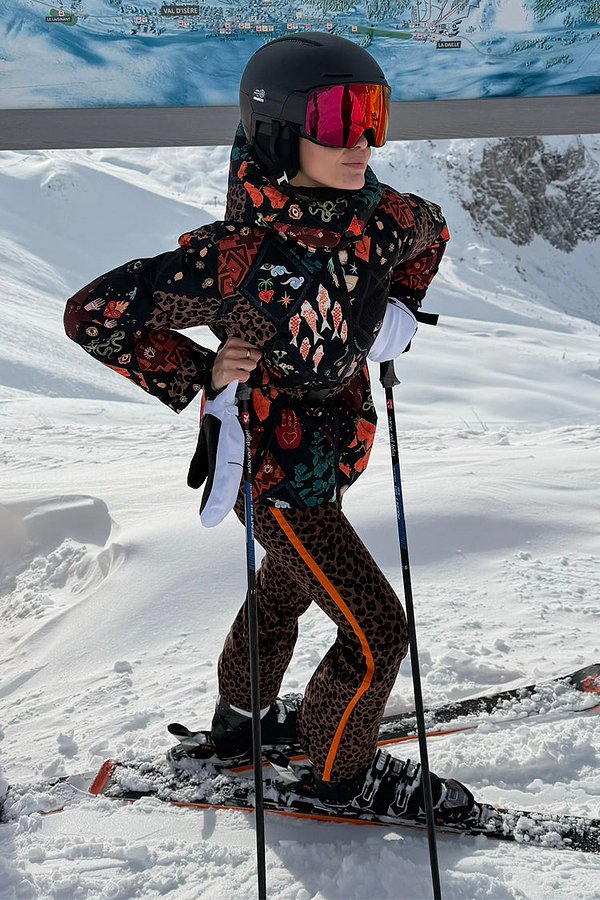 Leopard Ski Trousers with an orange side stripe, worn by a model on a snowy mountain with skis and a black helmet.