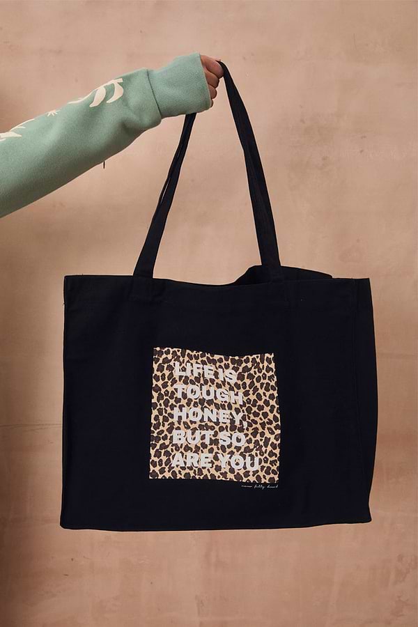 Black canvas Life Is Tough Tote Bag featuring a leopard print patch and motivational slogan, held against a beige wall.