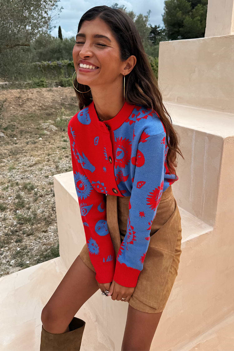 Blue and Red Holly Cardigan with sun and eye motifs and tortoise shell buttons, paired with a tan suede mini skirt.