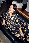 Deloris Satin Robe and pyjama trousers with iconic gold motifs, styled for lounging on a leather sofa.