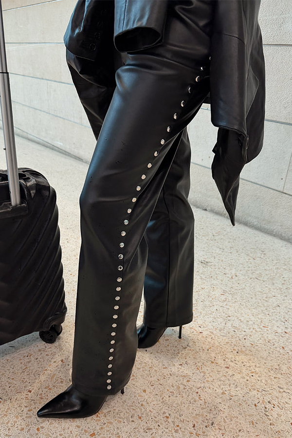 Black vegan leather studded leg trousers featuring silver side studs, styled with a suitcase for a vacation.