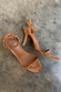 Tan suede stiletto heels with gold stud detailing and wrap-around ankle straps shown on a grey textured floor.