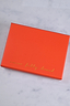 Vibrant orange rectangular jewellery box with gold Never Fully Dressed script on a white marble background.