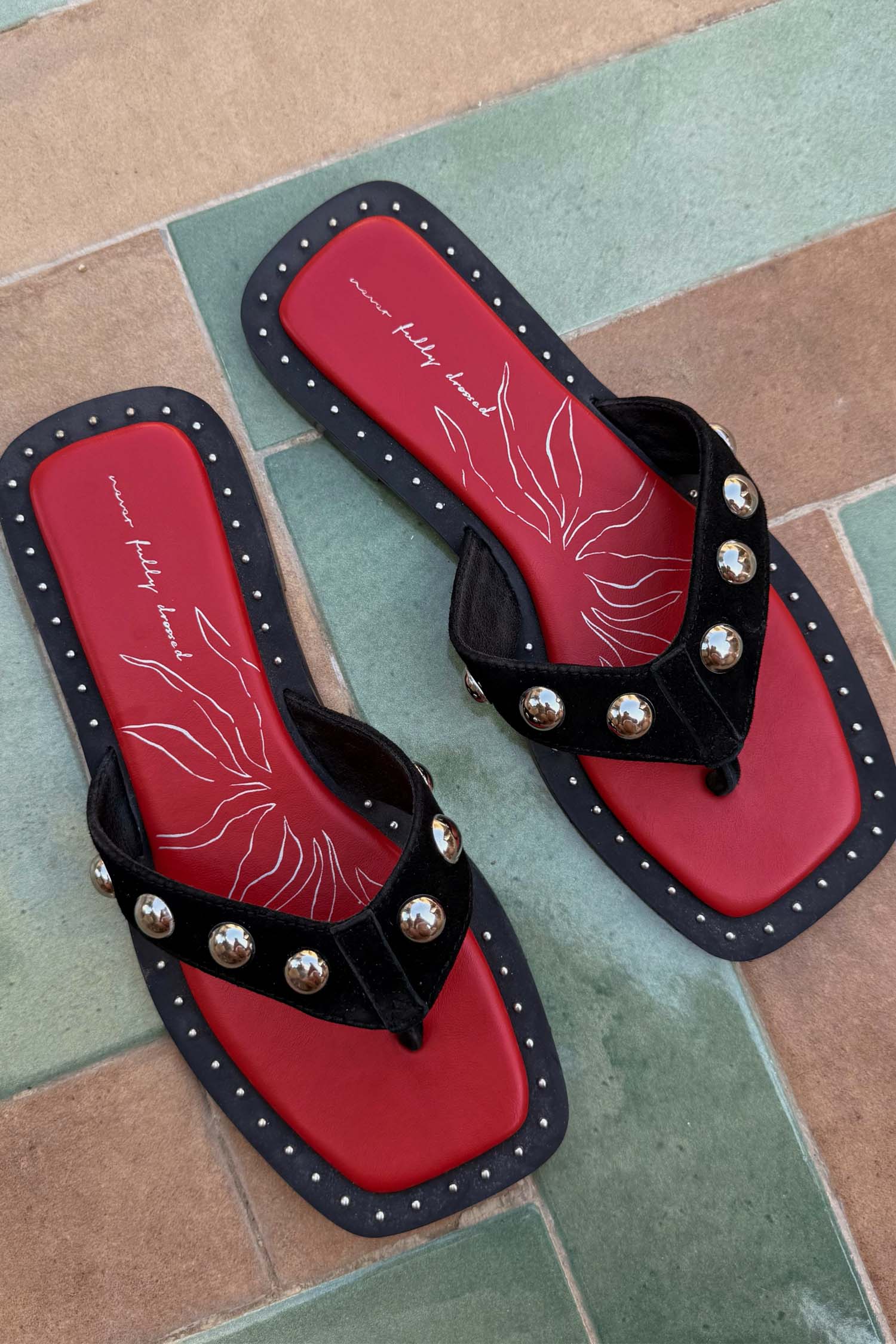Black suede flat sandals featuring silver studs, a square toe, and a red logo footbed with white graphic details.