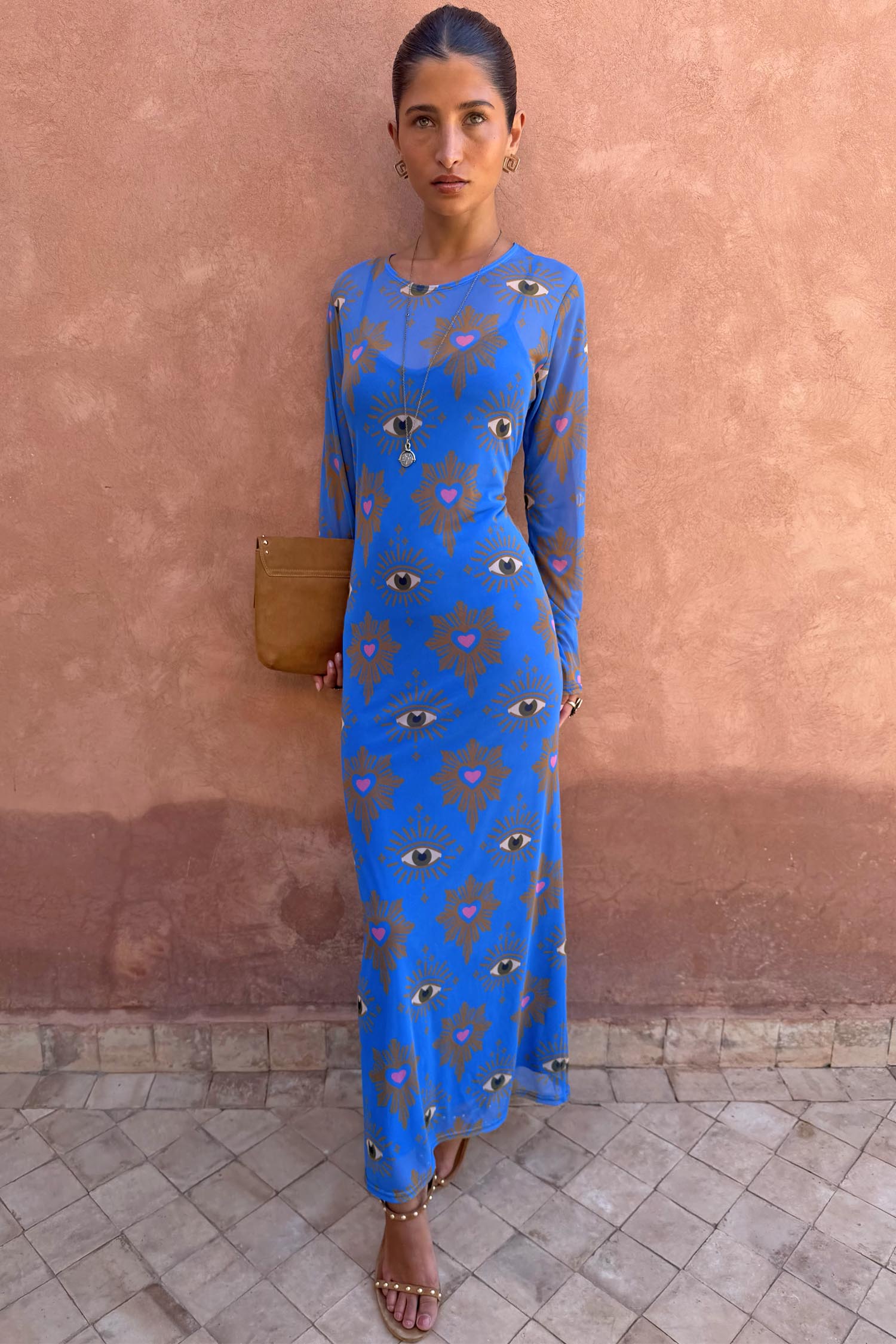 Blue Eye Mesh Maxi Dress with pink heart and eye motifs and long sleeves, worn by a model against a terracotta wall.