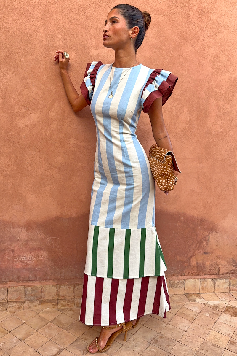 Blue Stripe Alena Midi Dress in cotton-linen with burgundy ruffle sleeves and a tiered hem against a terracotta wall.