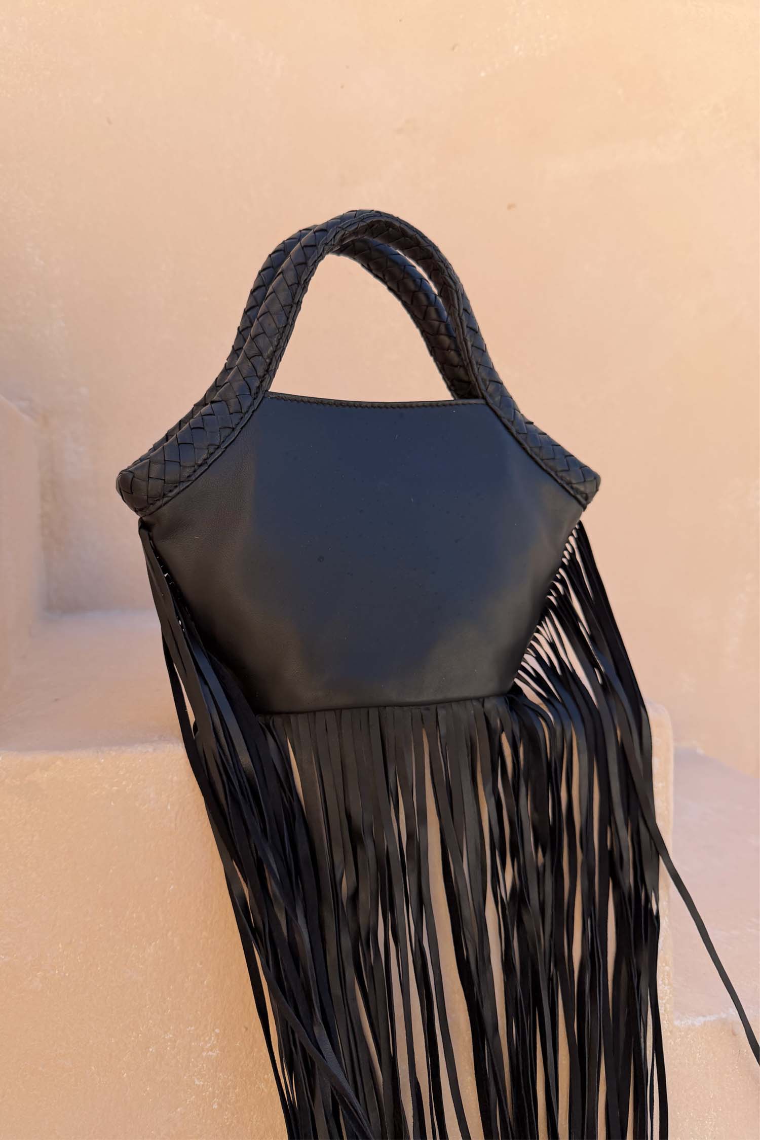 Black Leather Fringe Bag with a plaited handle and long fringe details, resting on a neutral stone background.