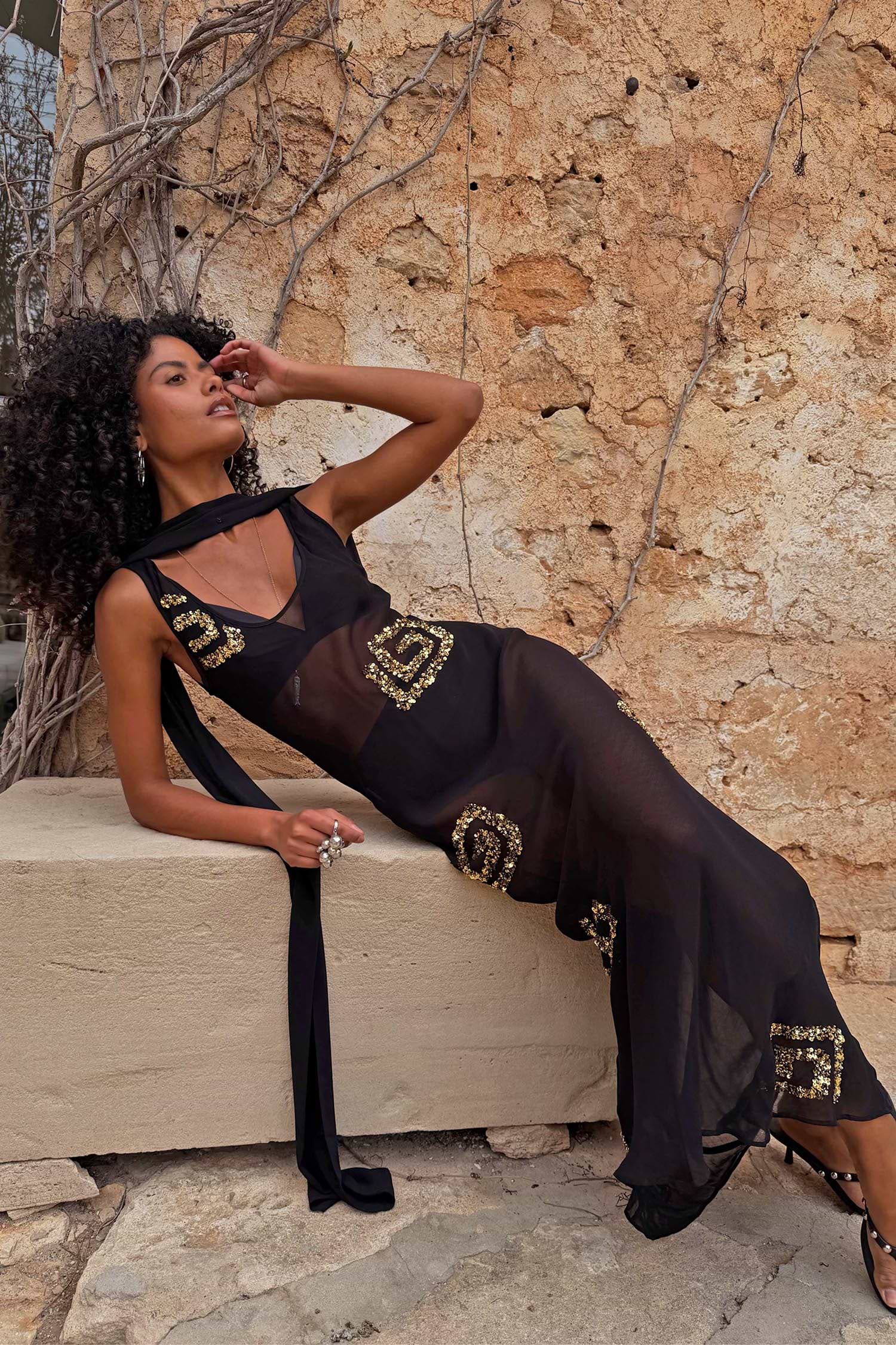 Petite Black & Gold Simona Maxi Dress in sheer mesh with gold sequins and a multi-wear scarf, styled against a stone wall.