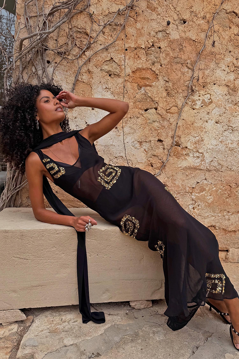 Petite Black & Gold Simona Maxi Dress in sheer mesh with gold sequins and a multi-wear scarf, styled against a stone wall.