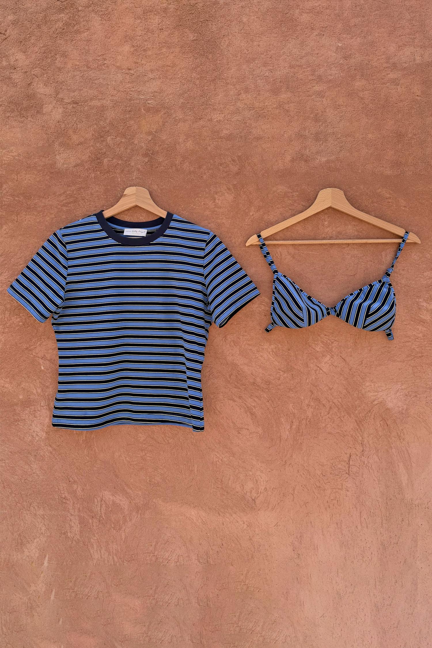 Blue Stripe Bra Baby T-Shirt with a detachable bra overlay, hanging on wooden hangers against a textured wall.