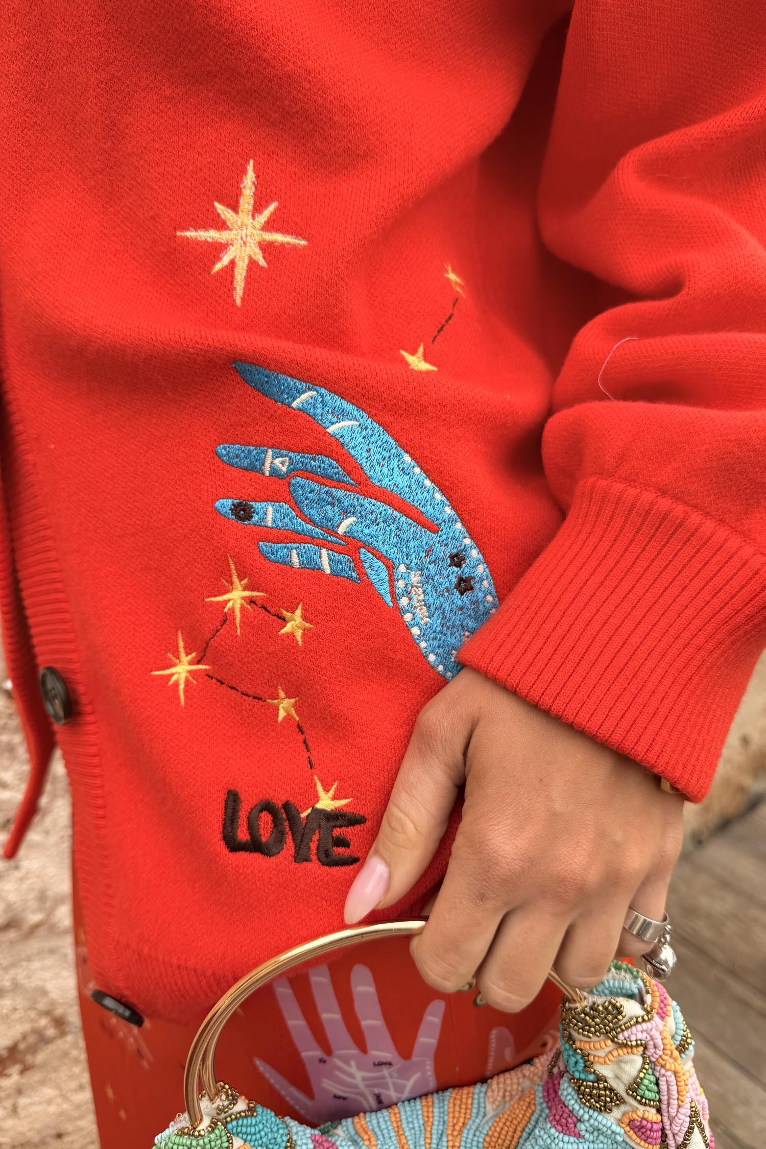 Red Celestial Cardigan detail with embroidered blue hand, yellow stars, and LOVE motif on a soft red knit base.