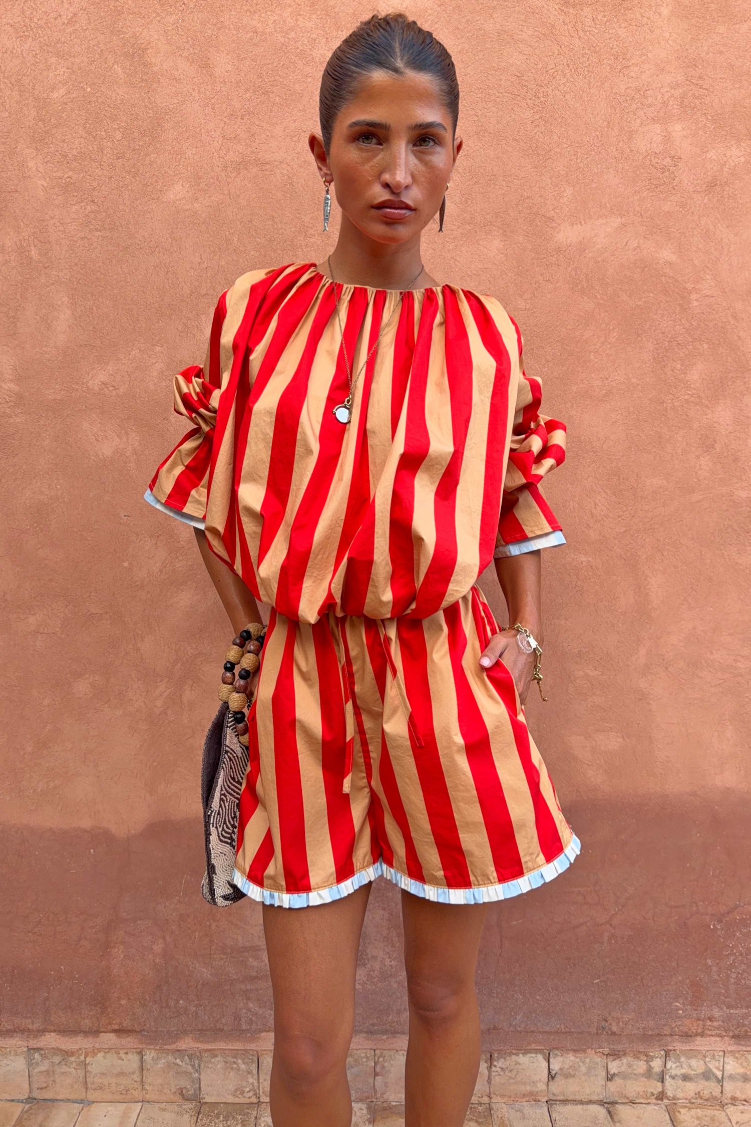 Tomato Stripe Ellie Shorts in red and camel cotton co-ord with contrast hem trim against a textured wall.