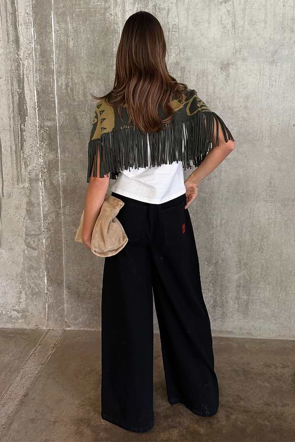 Olive Amara Knit Cape with tassel fringe and printed motifs, seen from the back with black wide-leg trousers.