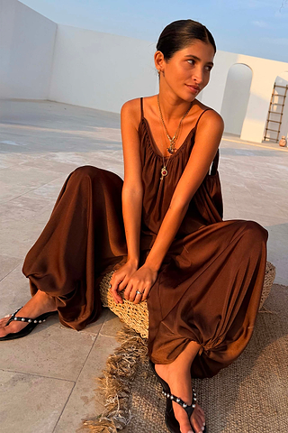 Chocolate Balloon Jumpsuit with a draped silhouette and gathered neckline, shown on a model sitting on a woven stool.