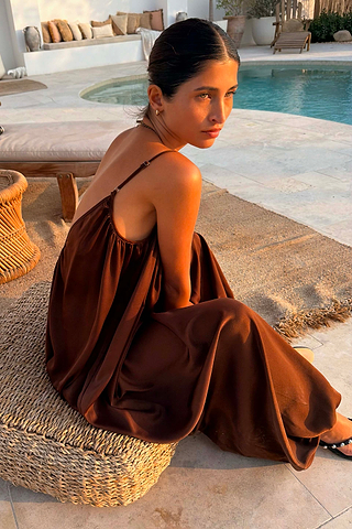 Chocolate Balloon Jumpsuit with a loose silhouette and deep V-back, worn by a model sitting on a woven stool poolside.
